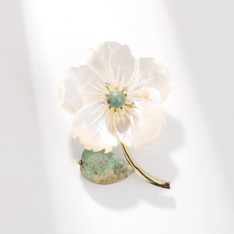 A delicate flower-shaped brooch with translucent white petals, a green stone center, and a green textured stem set in gold-tone metal, photographed on a clean white background with soft shadows.