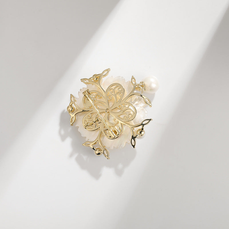 A gold-toned filigree flower brooch with a white pearl accent, displayed on a clean white surface with soft diagonal shadows.