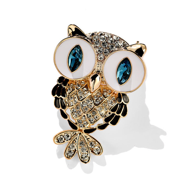 Owl-shaped brooch with gold-tone metal, white enamel eyes, blue gemstone eyes, and clear rhinestone detailing on a white background.