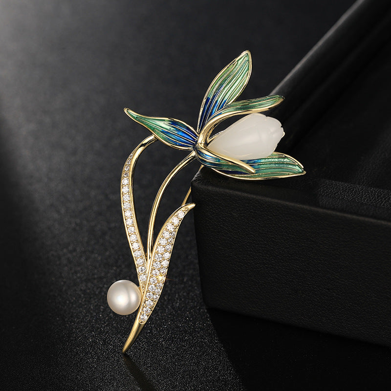 A gold-tone floral brooch featuring green and blue enamel leaves, a white pearl flower center, and a dangling white pearl bead, displayed on a black textured surface next to a black jewelry box.