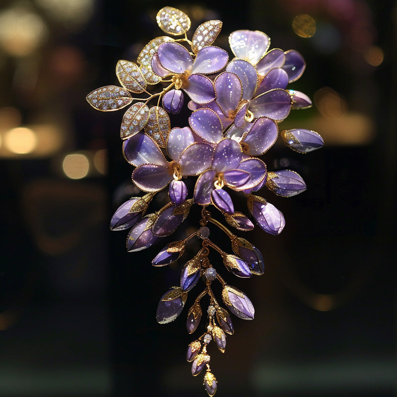 A purple wisteria flower brooch with gold-plated metal details, set with small clear crystals on the leaves and petals, shown against a dark, softly blurred background.