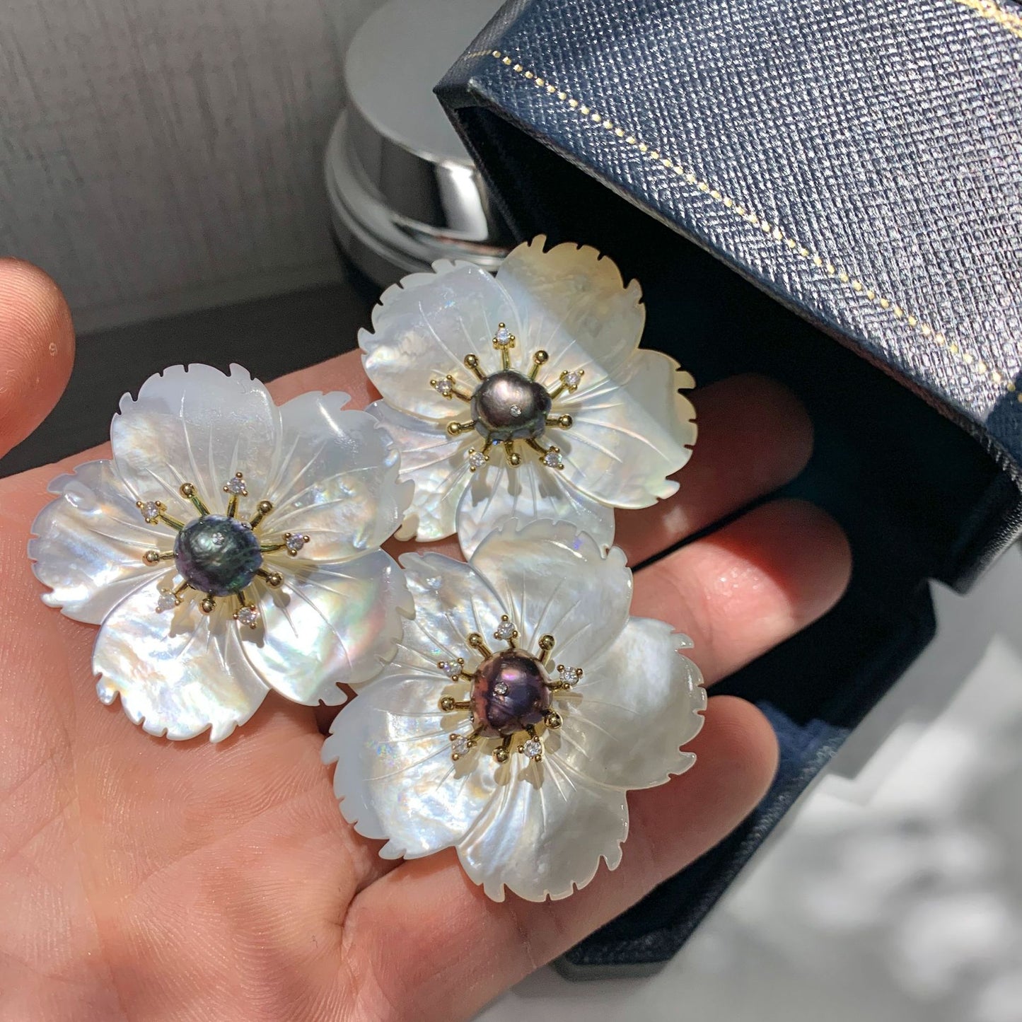 Three flower-shaped pearl earrings with iridescent white mother-of-pearl petals and gold-tone centers, held in a person's hand next to a textured dark blue jewelry box.
