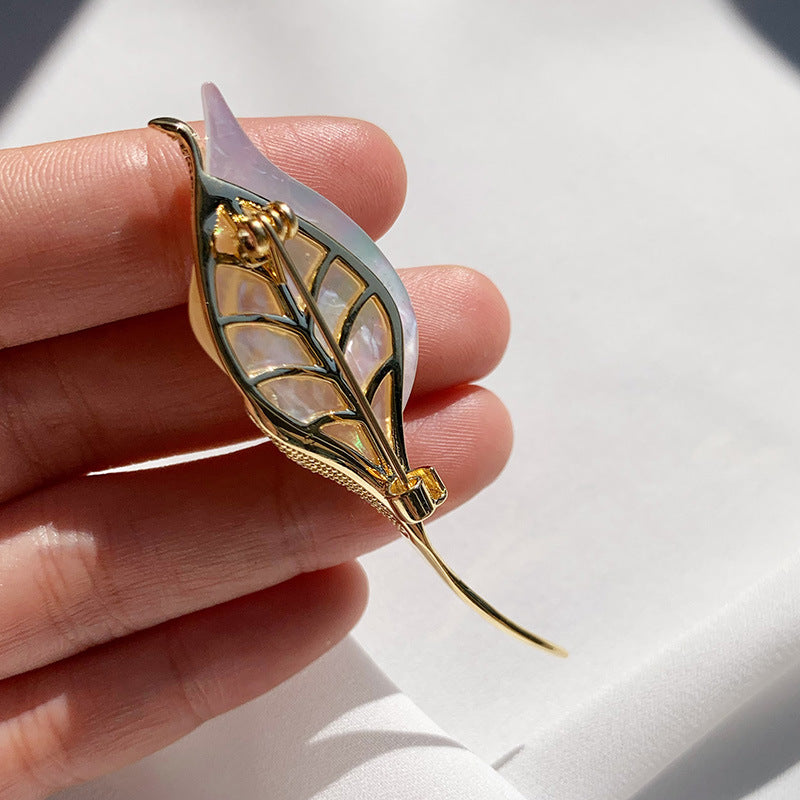 A gold-toned leaf-shaped brooch with iridescent mother-of-pearl inlay and a pale lavender tip, held between fingers against a white background.