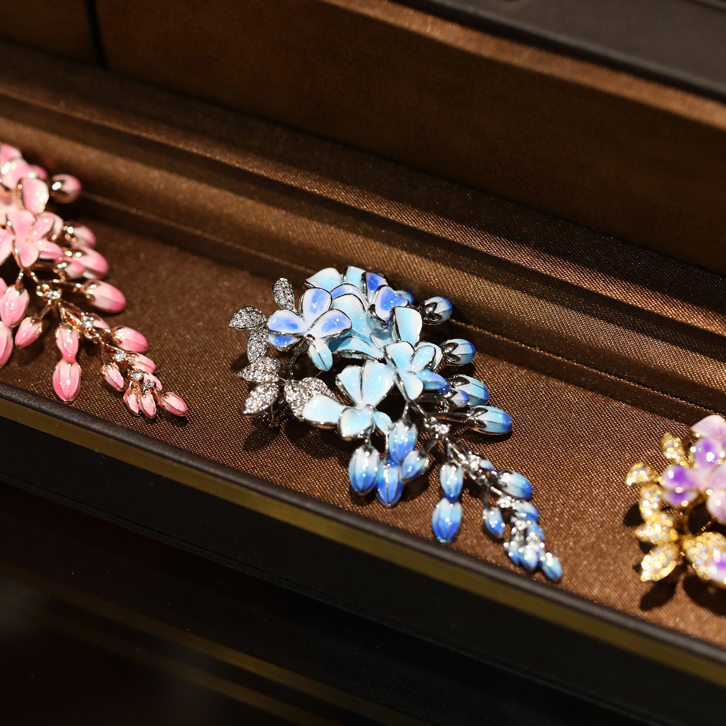 A close-up of a floral brooch with iridescent blue and white flower petals, silver-toned leaves, and small clear rhinestone accents, displayed on a brown velvet jewelry box. Partial pink and purple floral brooches are visible at the left and right edges.