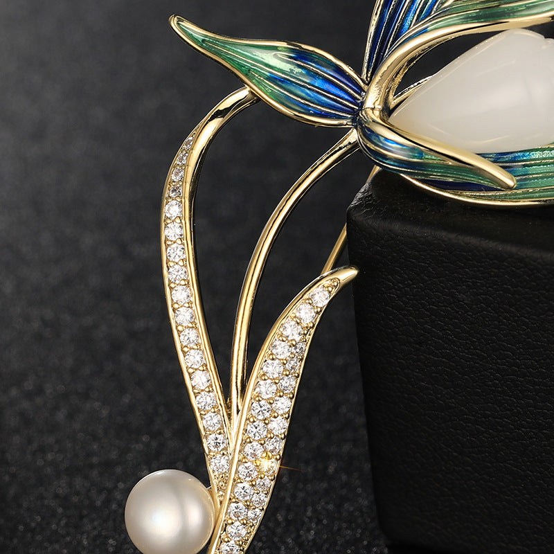 Gold floral brooch with green and blue enamel leaves, a white pearl bead, and clear crystal accents, displayed on a black jewelry box against a dark background.