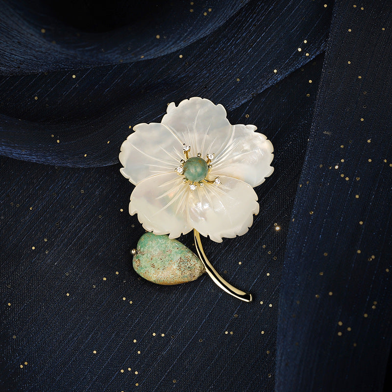 A delicate flower-shaped brooch with translucent white petals, a green stone center, and a green-and-gold leaf, displayed on a dark navy textured fabric background.