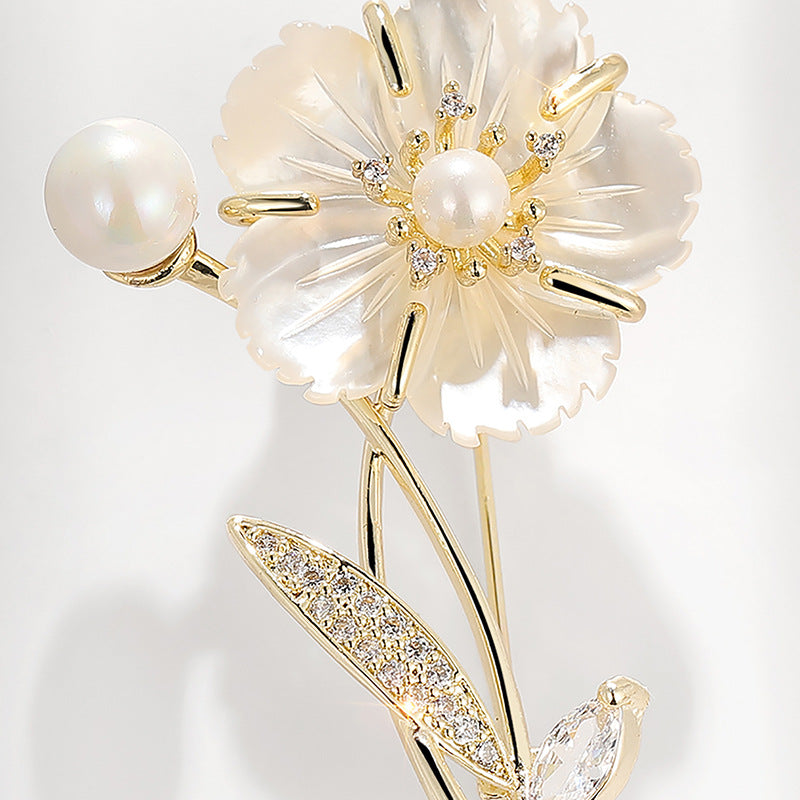 A close-up of a floral brooch featuring a large cream-colored mother-of-pearl flower with a pearl center, gold-tone metal accents, and small clear stones, alongside a pearl bead and a leaf-shaped element set with clear crystals, on a light gray background.