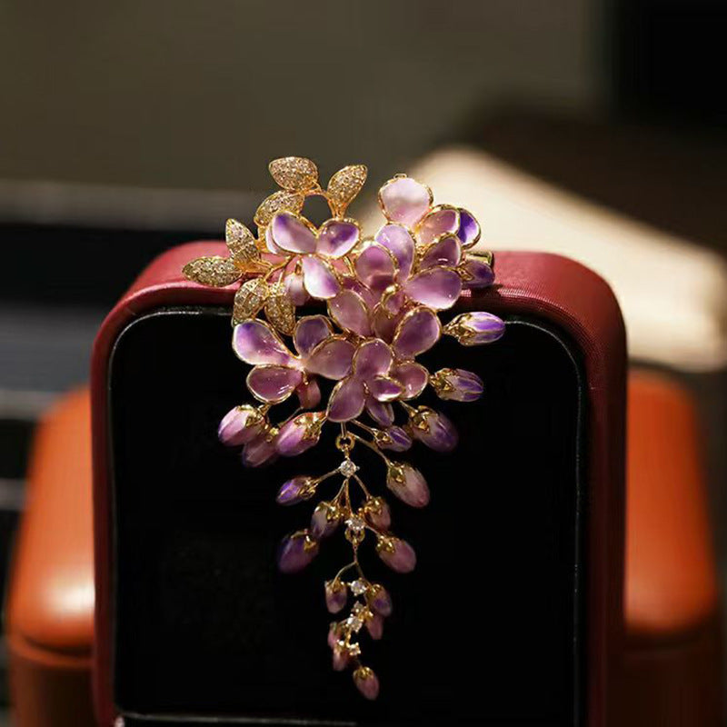 A floral brooch with lavender-purple petals and gold-toned leaves, displayed on the edge of an open dark red jewelry box with a black interior.