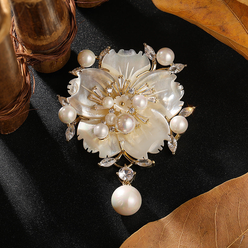 A floral pearl brooch with gold-tone metal detailing, set on a black textured background with decorative leaves and copper-toned objects.