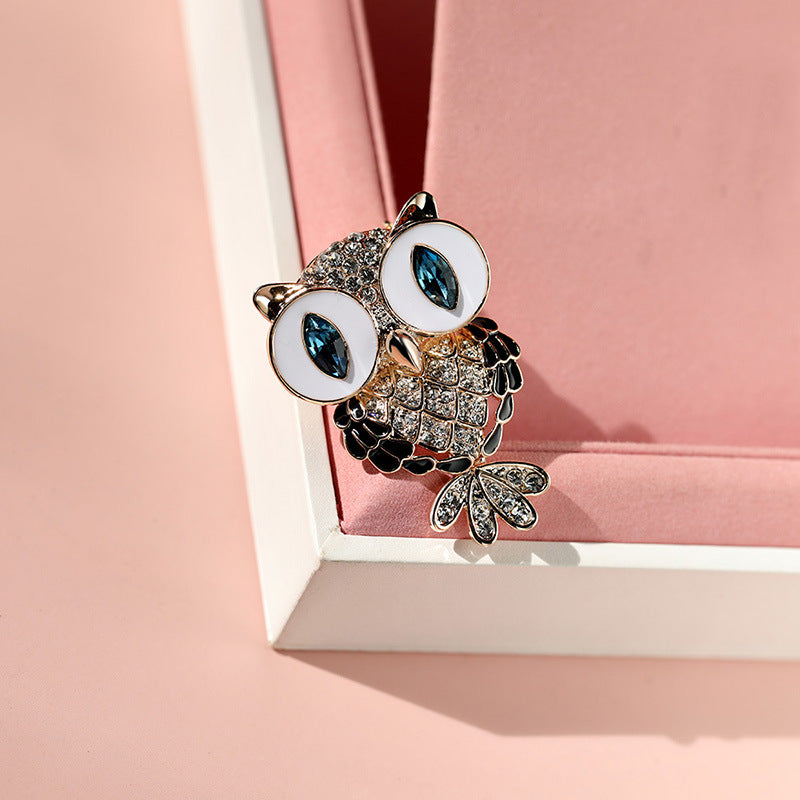 An owl-shaped brooch with large white oval eyes, dark blue marquise-cut stones, and a body covered in small clear stones, displayed on a pink jewelry box corner against a pink background.