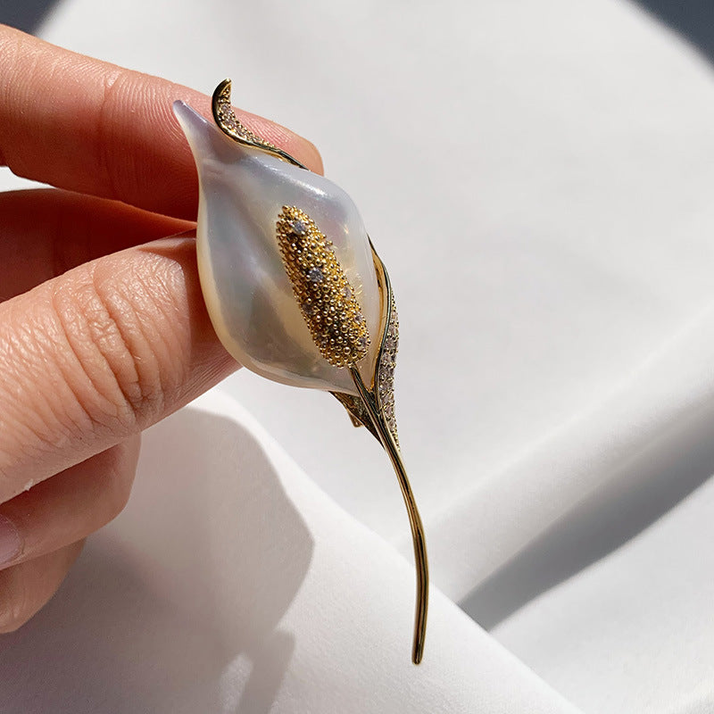 A close-up of a hand holding a calla lily flower brooch. The brooch features a translucent white mother-of-pearl bloom with a textured gold center and gold leaf-like accents, set against a soft white draped fabric background.