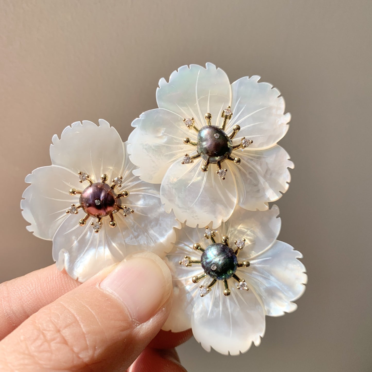 Three white flower-shaped brooches with iridescent centers and gold-toned metal accents, held between fingers against a plain beige background.