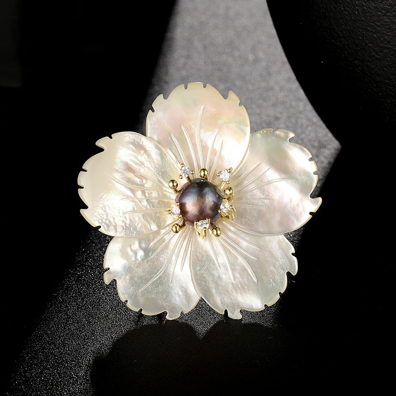 A flower-shaped brooch featuring iridescent white mother-of-pearl petals with gold-tone metal accents and a dark pearl center, displayed on a black background.