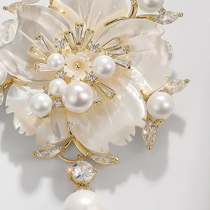 A close-up of a floral brooch featuring layered mother-of-pearl petals, faux pearls, and gold-tone metal with clear crystal accents on a light gray background.