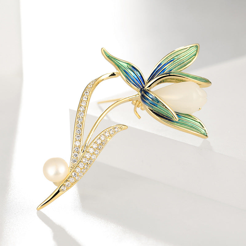 A gold-tone floral brooch featuring green and blue enamel petals, a white pearl center, and a curved leaf with small clear stones and a pearl accent, displayed on a white geometric surface.