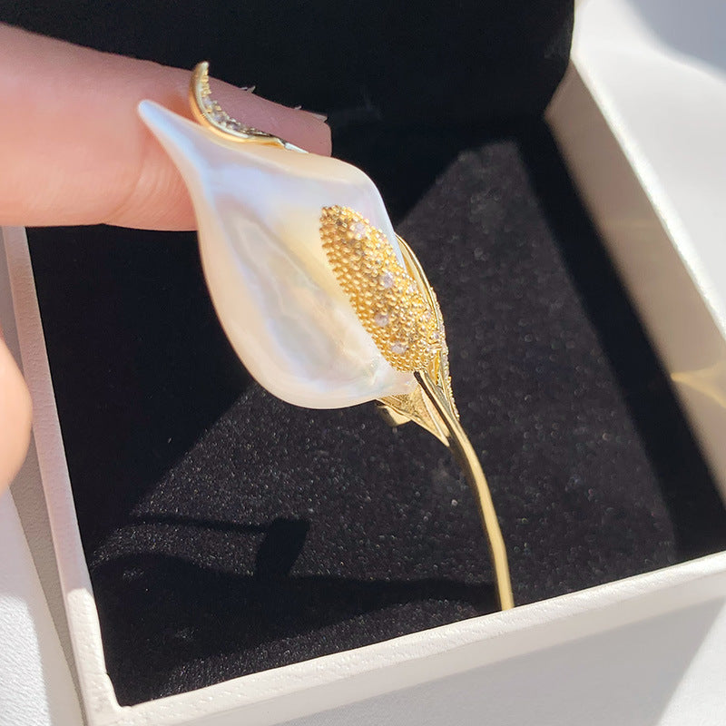 A gold calla lily hair claw clip with a white mother-of-pearl flower petal, held by a hand over a black velvet-lined jewelry box.