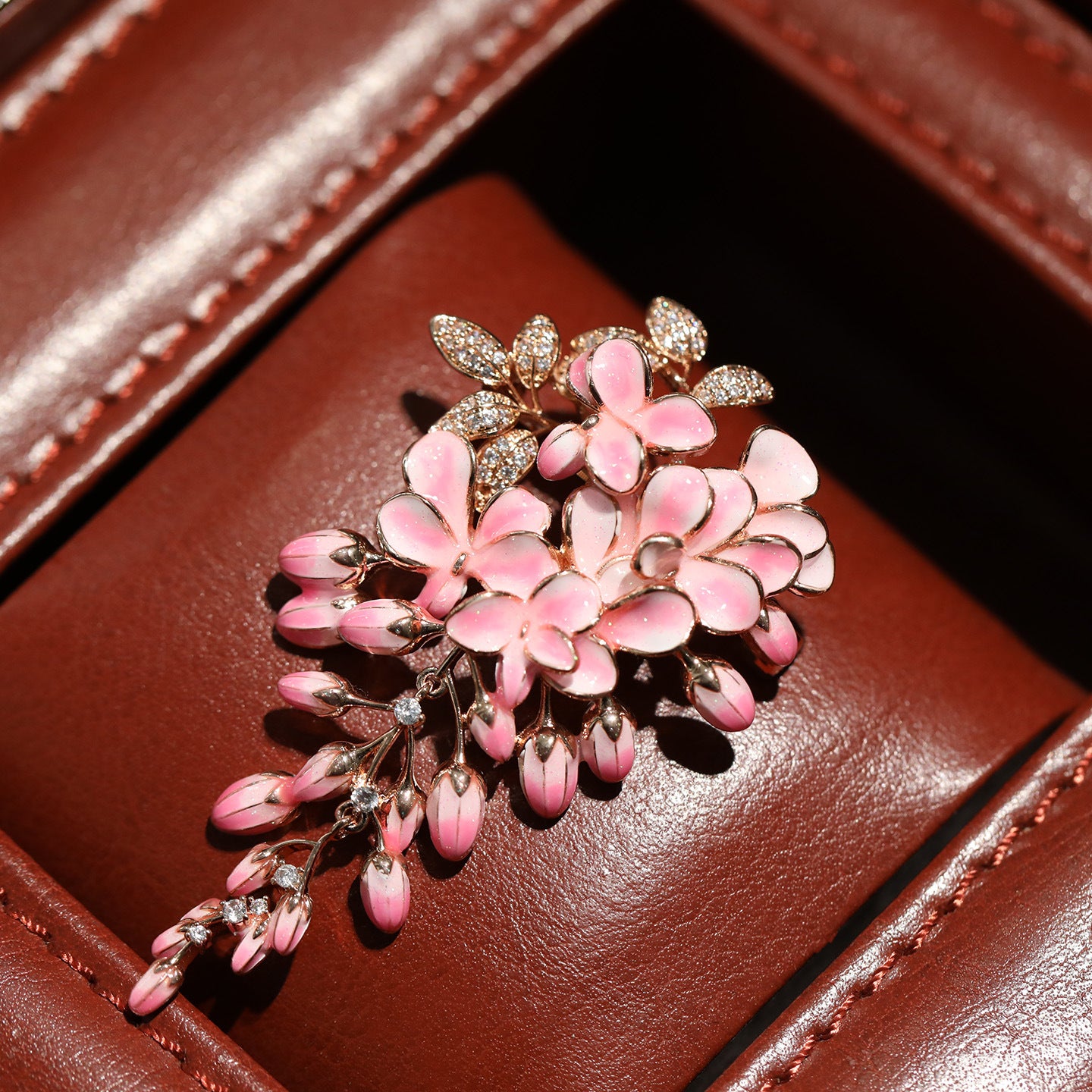 A close-up of a floral brooch with pink enamel flowers and gold-toned leaves, some accented with small clear stones, displayed on a brown leather surface.
