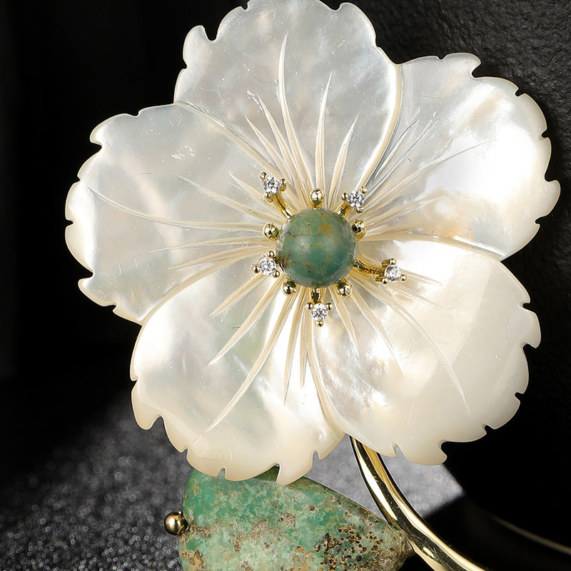 A close-up of a flower-shaped brooch or pin made from iridescent white mother-of-pearl petals with a central green stone and small clear stones in a gold-tone setting, shown against a dark background.