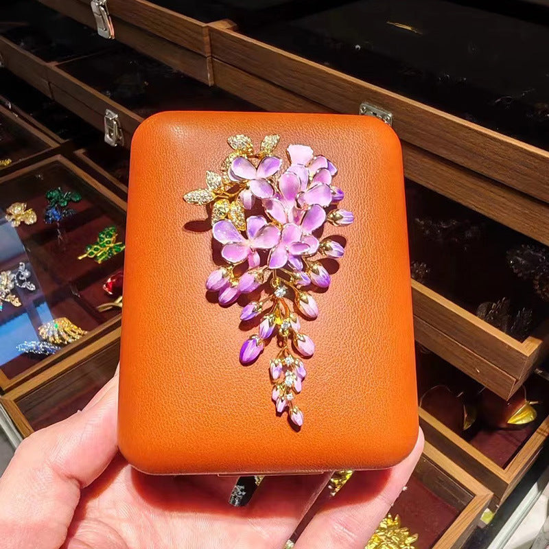 A hand holds a small orange leather clutch decorated with a large purple floral brooch featuring gold-tone metal and rhinestones, set against a background of wooden jewelry display cases.