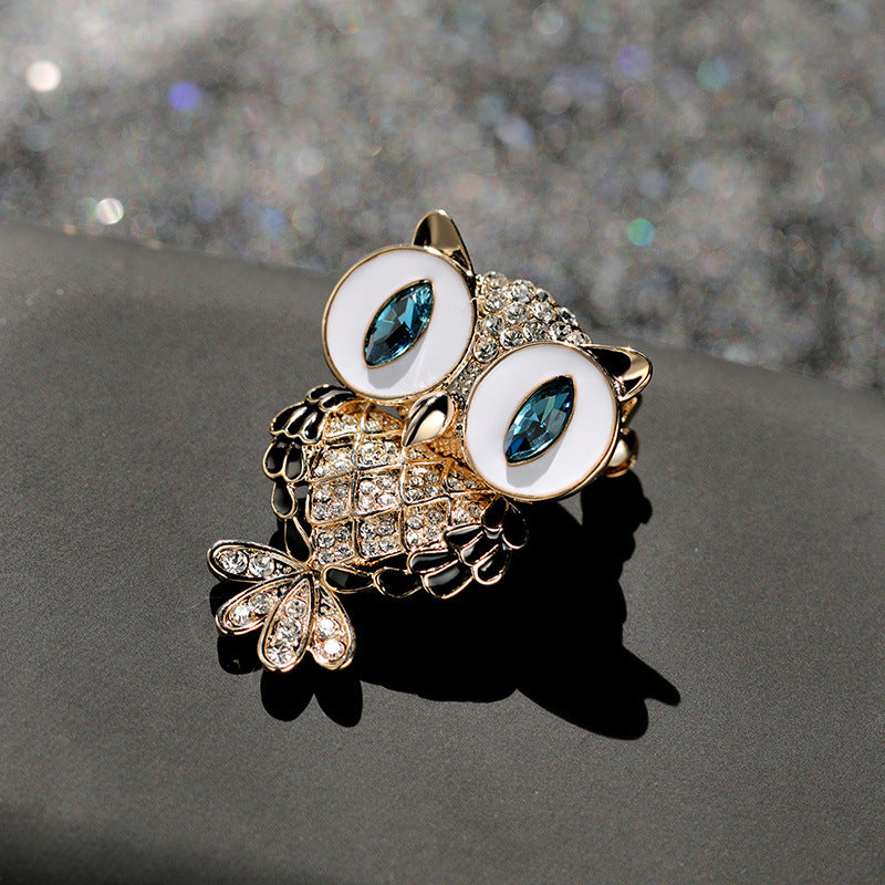 Owl-shaped ring with gold-tone metal, white enamel, and blue marquise stones in the eyes, accented with clear rhinestones, photographed on a dark surface with a glittery silver background.