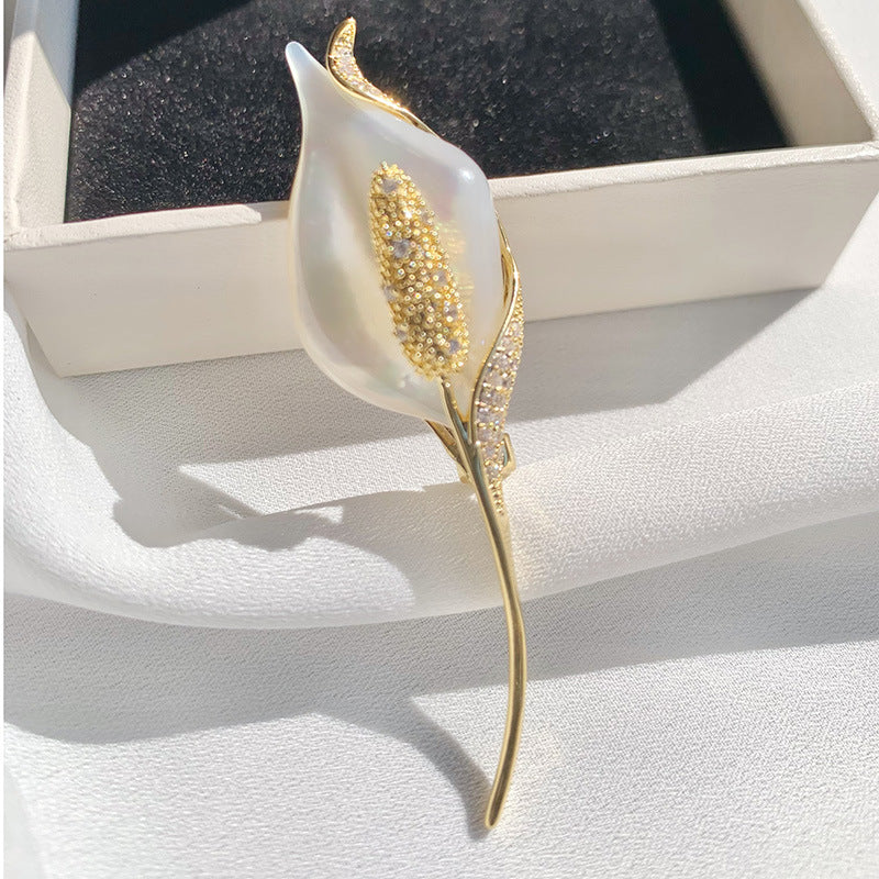 A calla lily flower brooch with a white faux-pearl center, gold textured center and leaf, and small clear crystals along the side, displayed on a white fabric surface next to its presentation box.
