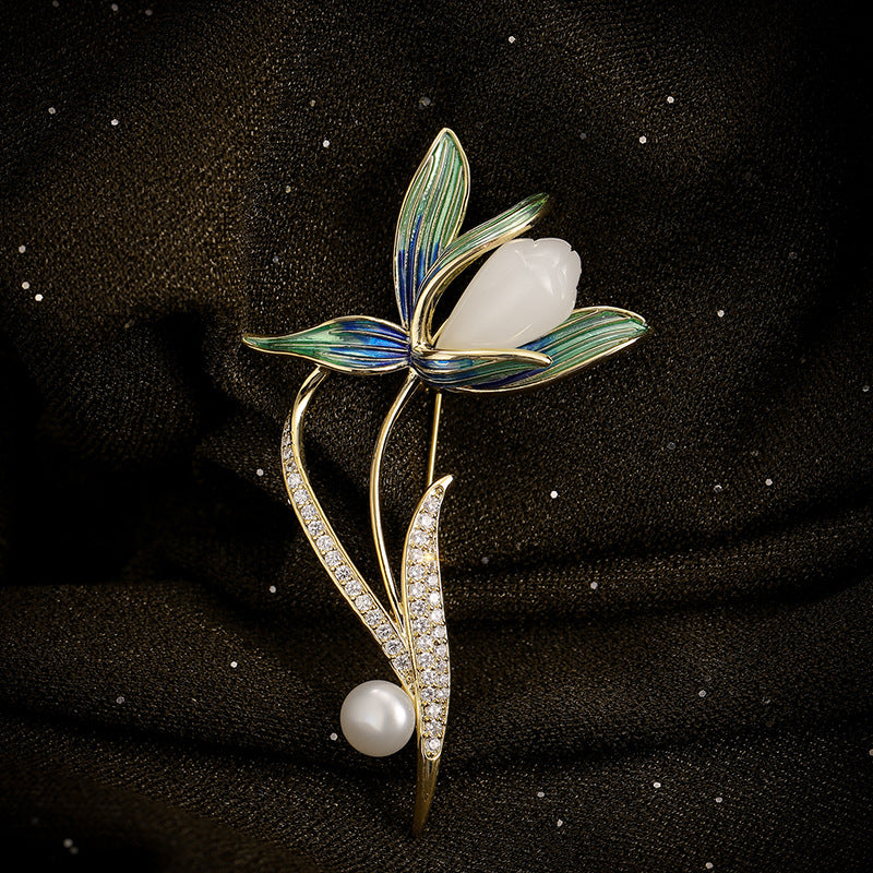 An elegant floral brooch featuring a white pearl flower, green and blue enamel leaves, and gold-toned stems with a pavé-set pearl leaf and a single white pearl, displayed on a textured black fabric background.