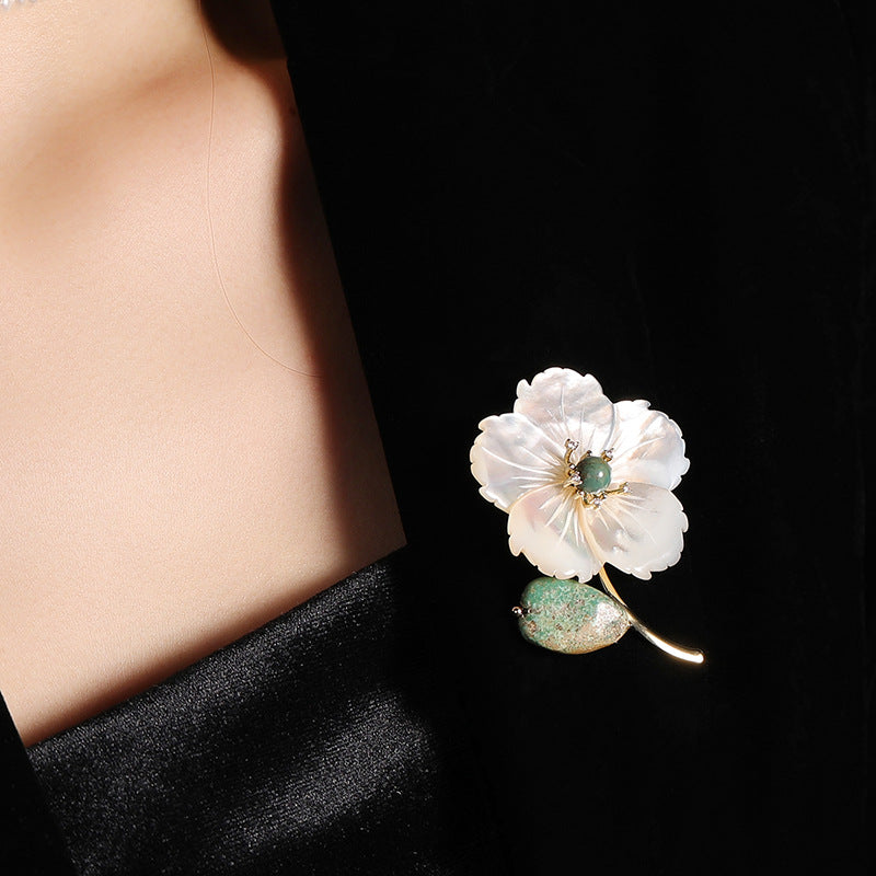 A close-up of a white flower brooch with green and gold details pinned to a black garment on a model's chest.