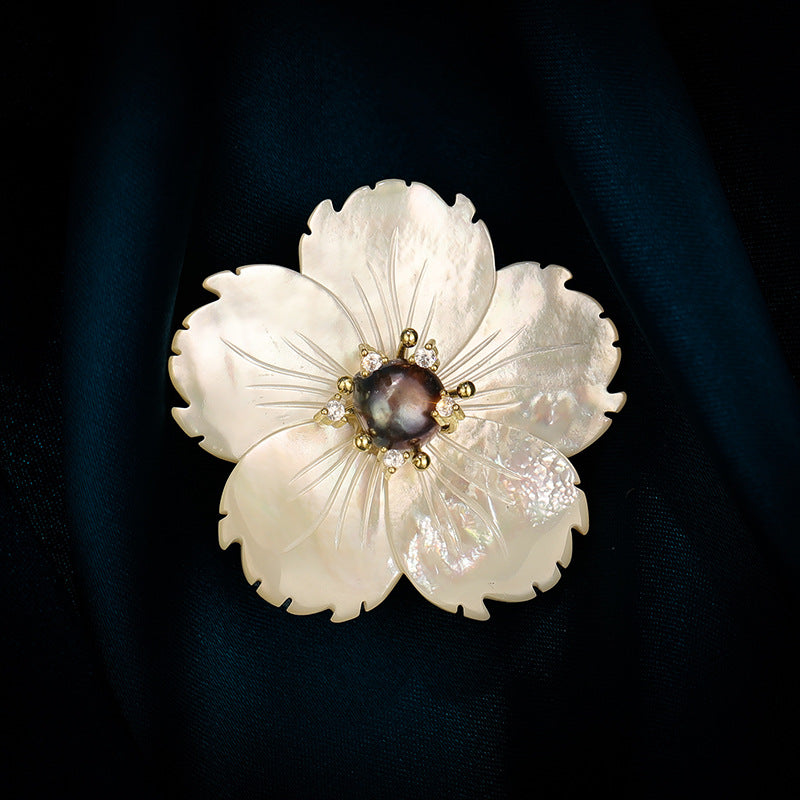 A white mother-of-pearl flower brooch with gold-toned metal accents and a dark pearl center, displayed on a dark navy blue satin fabric background.