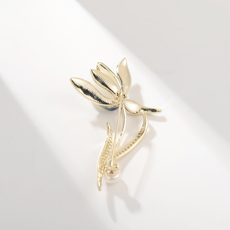 A gold-tone metal dragonfly brooch with textured wings and a curved stem, photographed on a clean white surface with soft shadows.