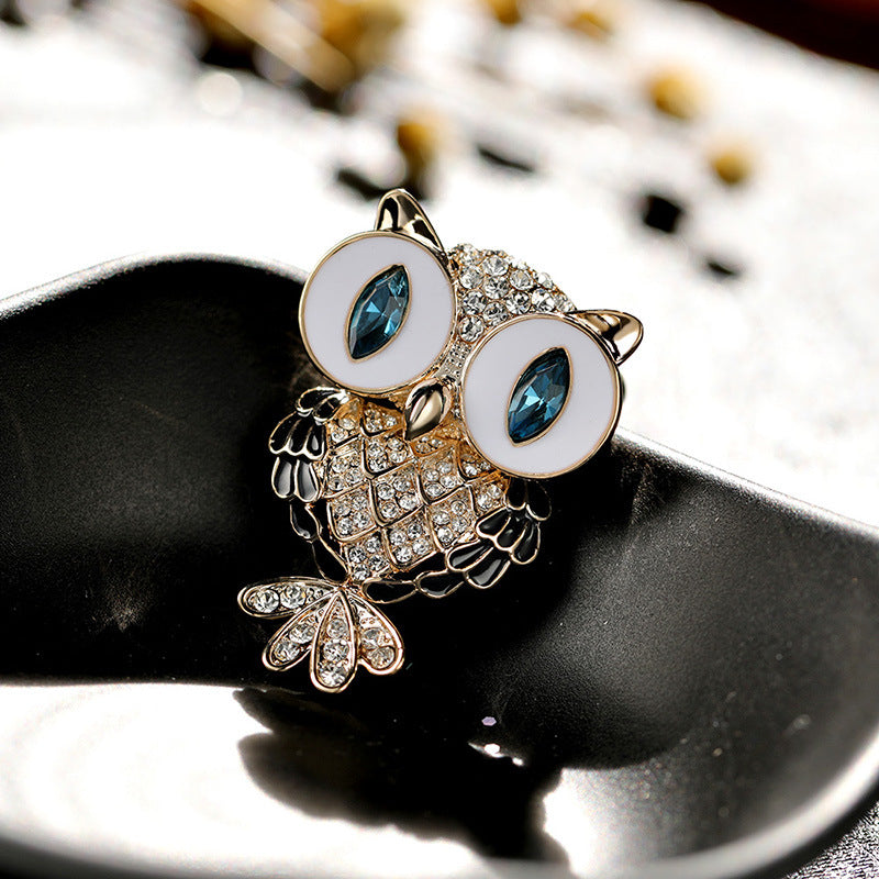 Owl-shaped brooch with white enamel eyes, blue marquise stones, and gold-tone crystal-embellished details, displayed on a black reflective surface.
