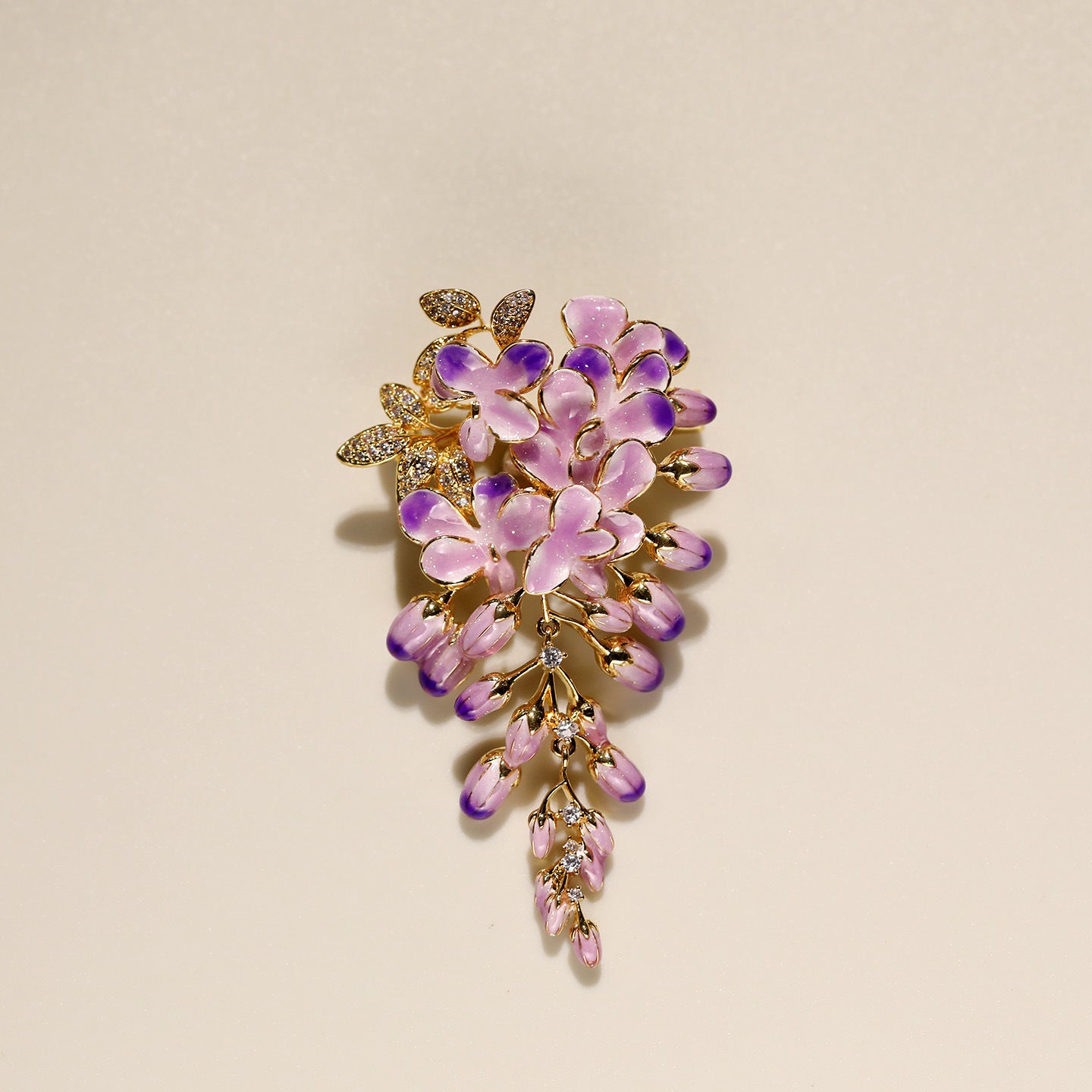 A gold-tone floral brooch featuring lavender and light pink enamel flowers with small clear rhinestone accents, displayed on a warm beige background.