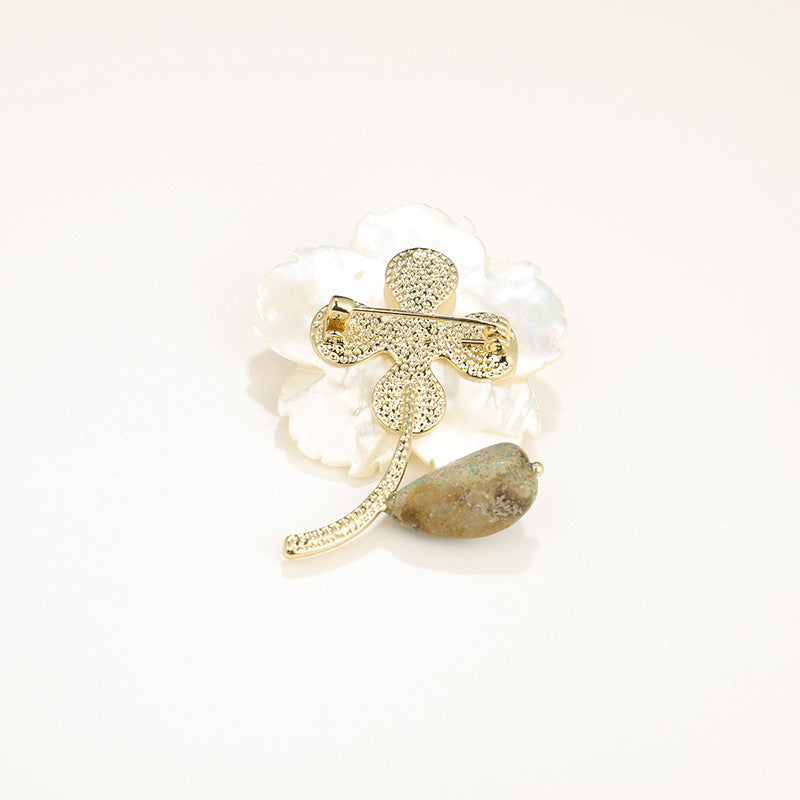 A gold-toned floral brooch featuring a textured cross, a curved stem with a small bead, and a white mother-of-pearl flower with a brown stone accent, set against a plain white background.