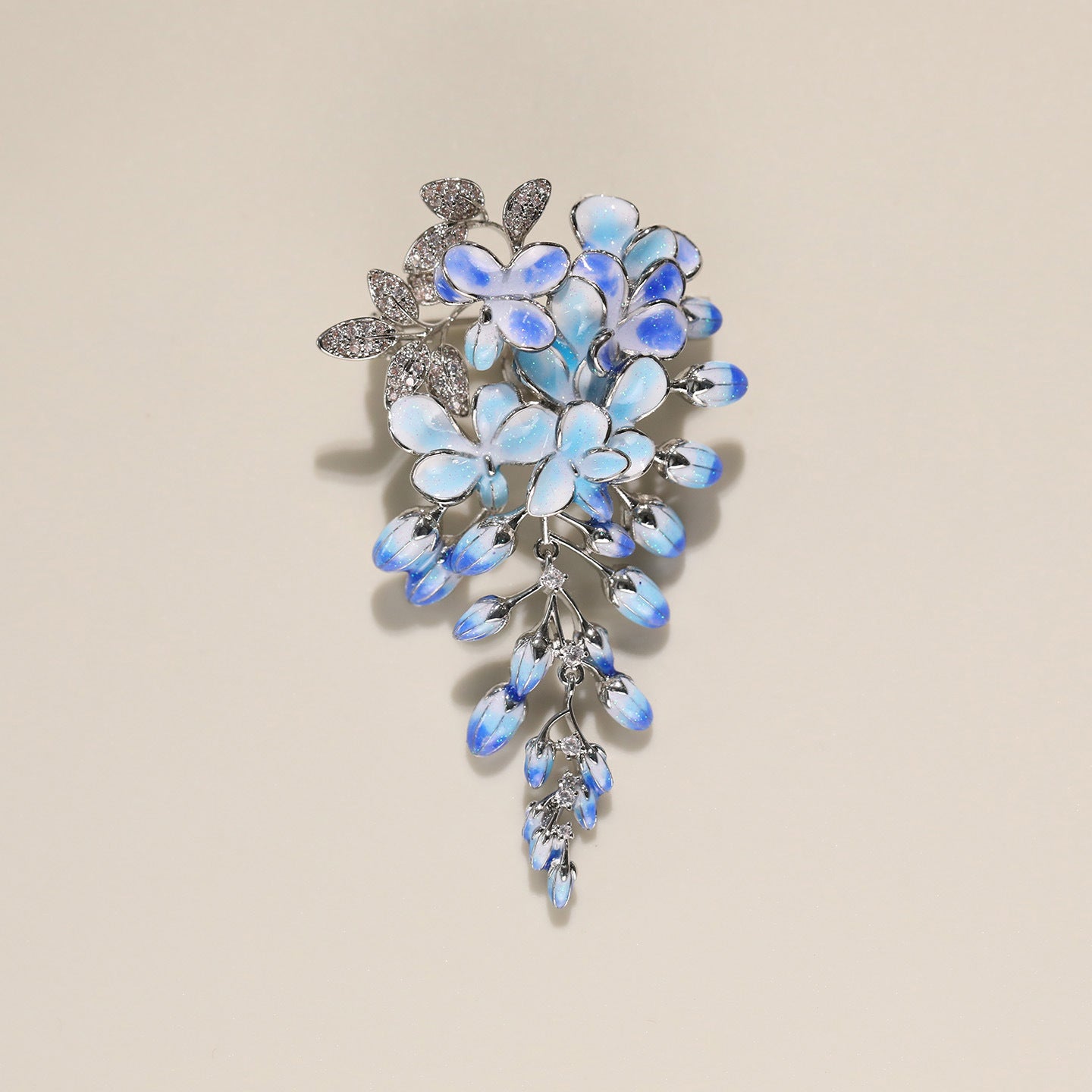 A floral brooch featuring light blue iridescent flowers and teardrop-shaped blue accents on a silver-toned setting, photographed against a solid beige background.