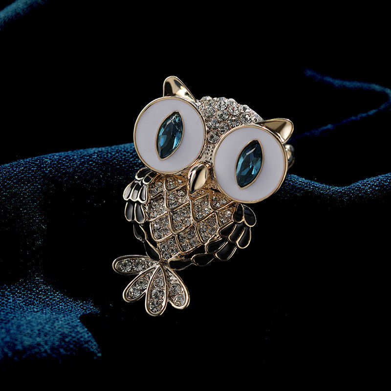 An owl-shaped brooch with a gold-tone metal body, white enamel around the eyes, and marquise-cut blue stones in the eyes, set against a dark blue velvet fabric background.