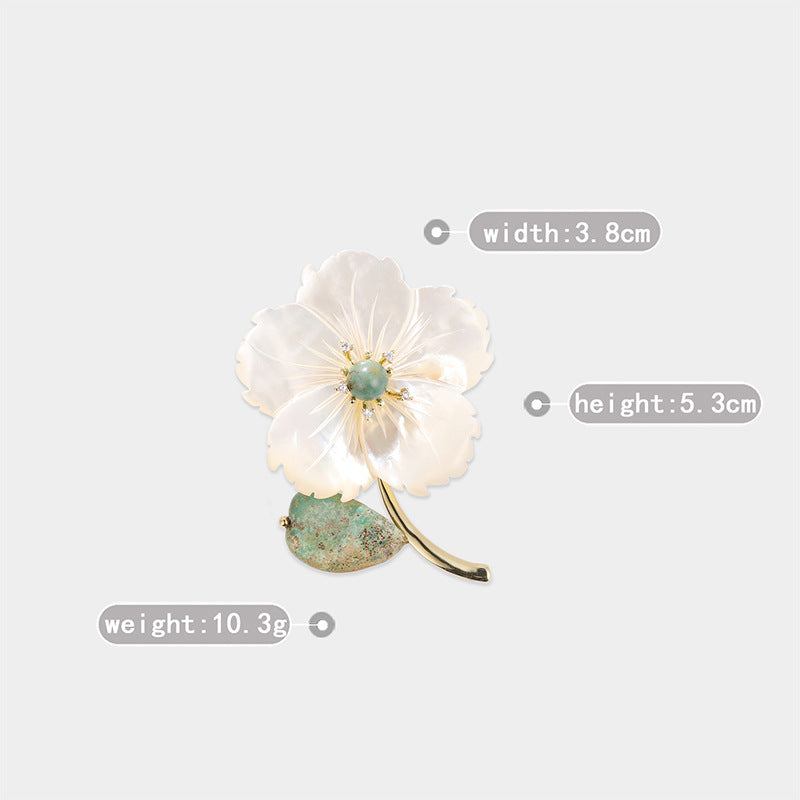 A flower-shaped brooch with white mother-of-pearl petals, a light green center, and a textured green leaf base on a gold-toned stem, shown on a light gray background with measurement labels for width, height, and weight.