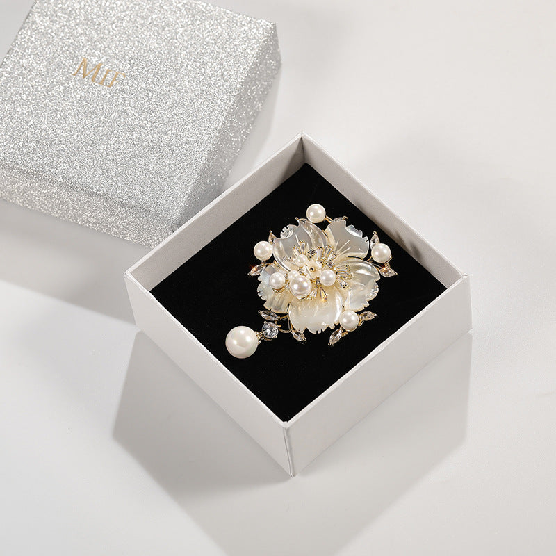 A floral pearl brooch with gold-tone metal and clear rhinestones, displayed in an open white jewelry box with a black velvet insert. The silver glitter lid with gold 'MD' lettering sits behind it on a clean white background.