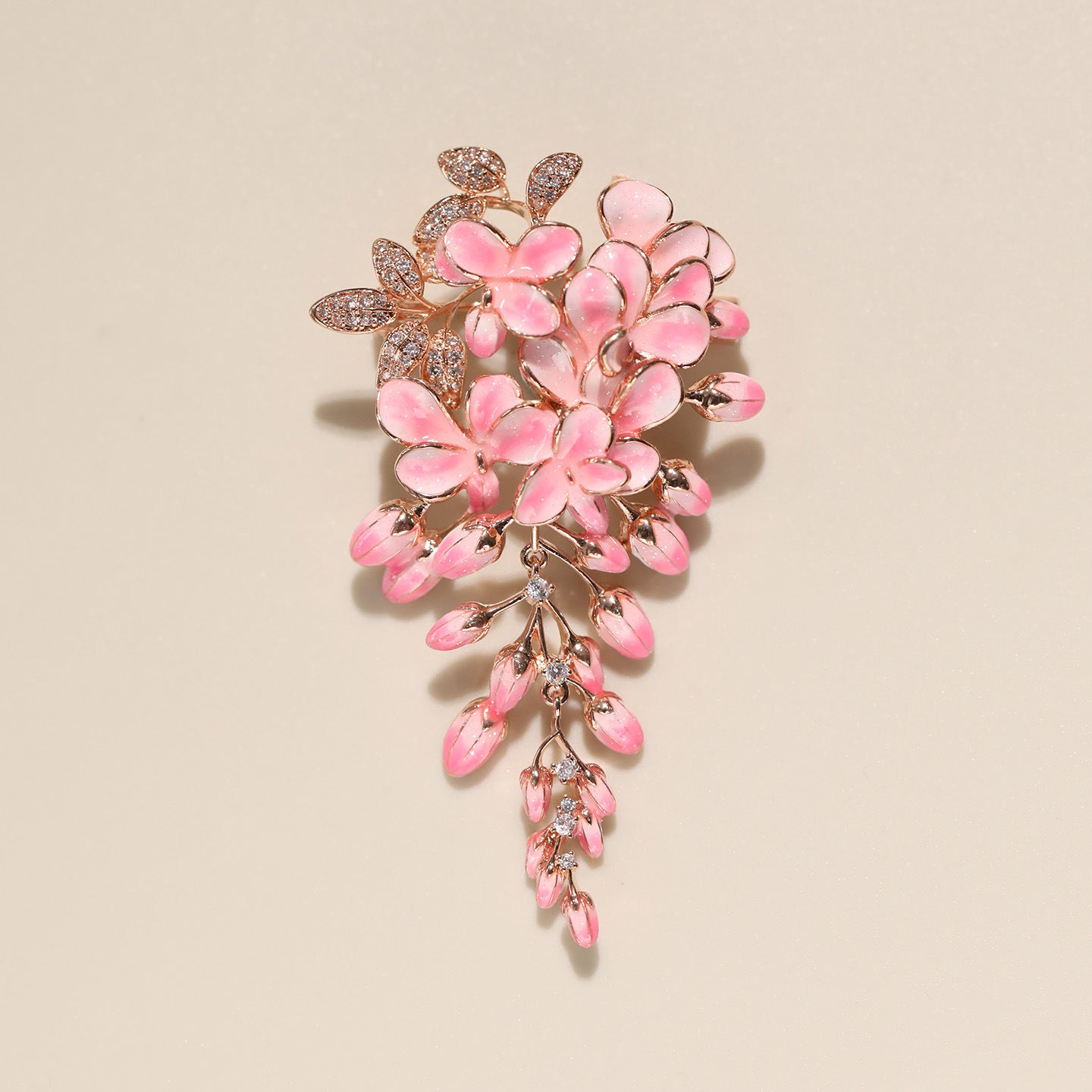A pink floral brooch featuring glossy pink enamel petals and leaves with sparkling clear crystals, set in a rose gold-tone metal against a solid beige background.