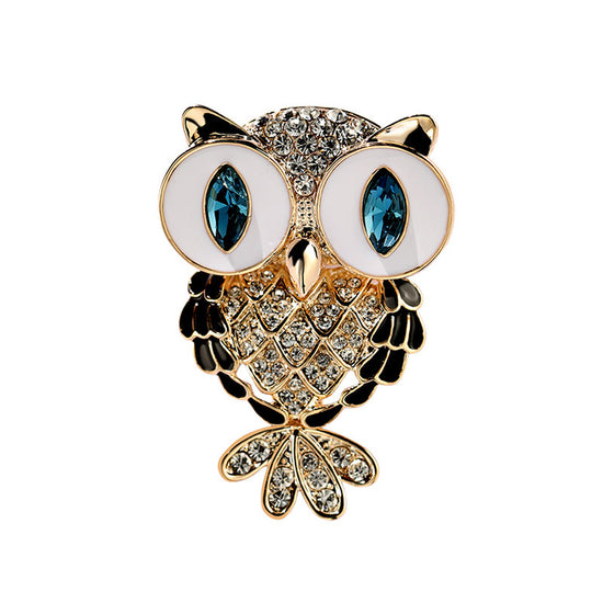Owl-shaped brooch with gold-tone metal, white enamel eyes, blue gemstone eyes, and clear rhinestone detailing on a plain white background.
