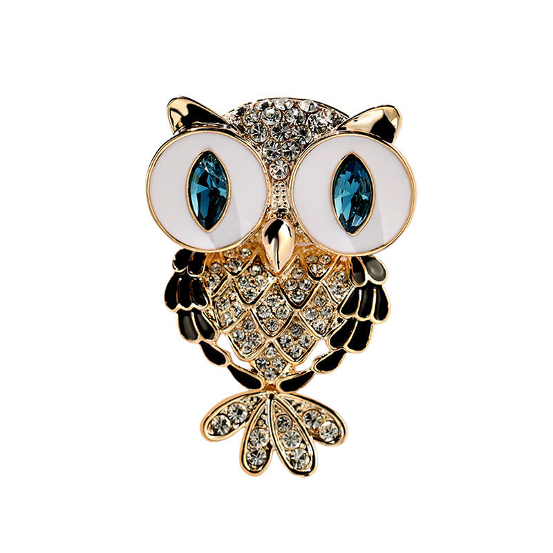 Owl-shaped brooch with gold-tone metal, white enamel eyes, blue gemstone eyes, and clear rhinestone detailing on a plain white background.
